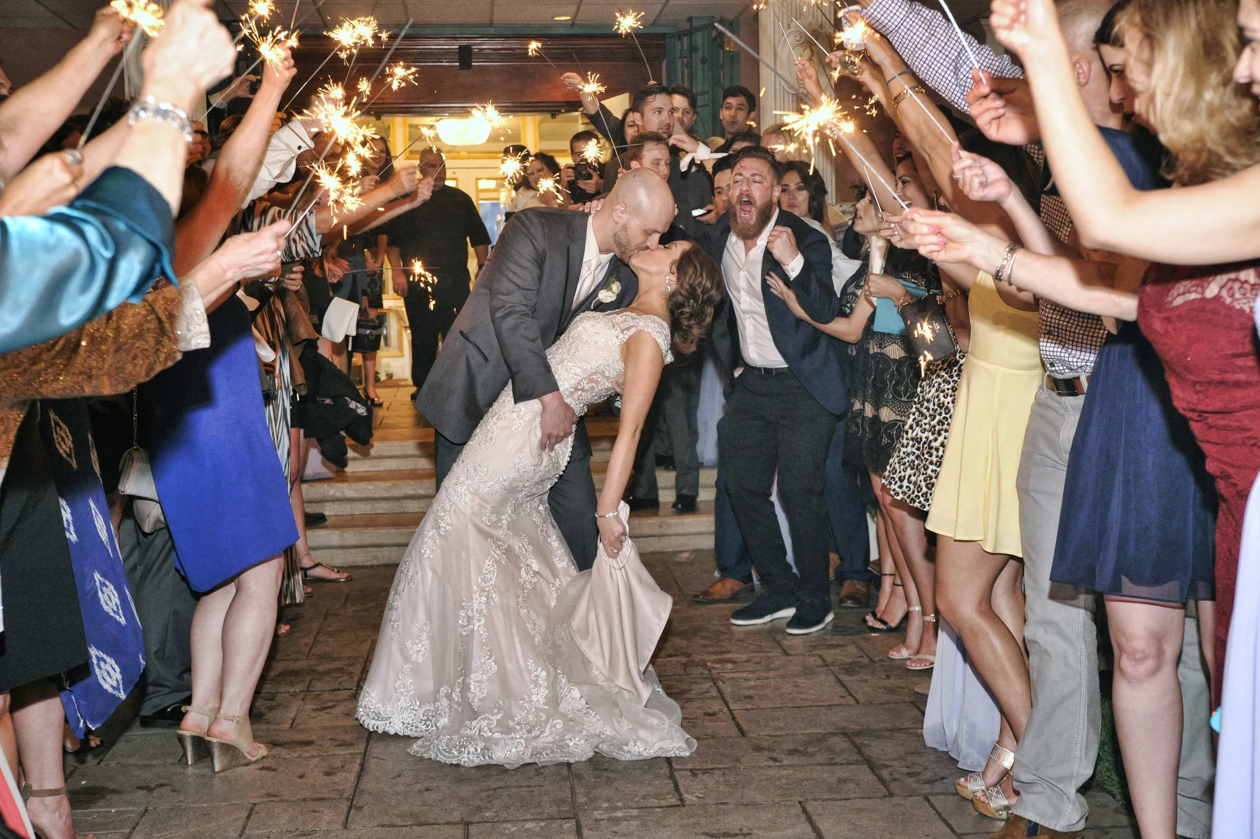 Bride and groom kissing in a dip pose surrounded by guests holding sparklers at a wedding celebration.