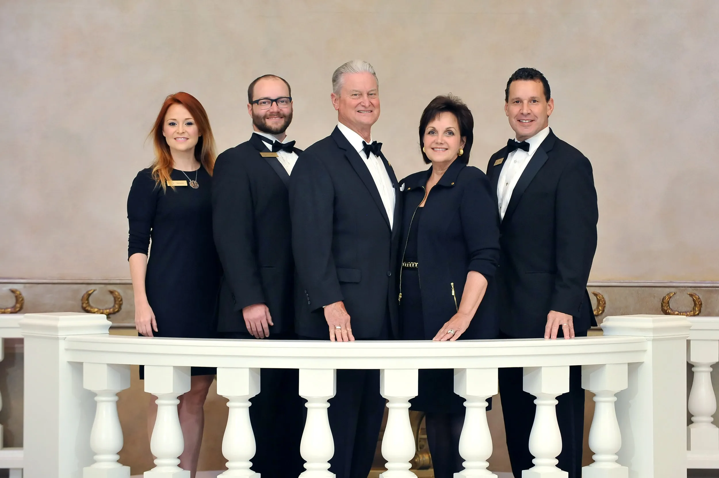 Group of five adults in formal black attire standing behind a white balcony railing, smiling.