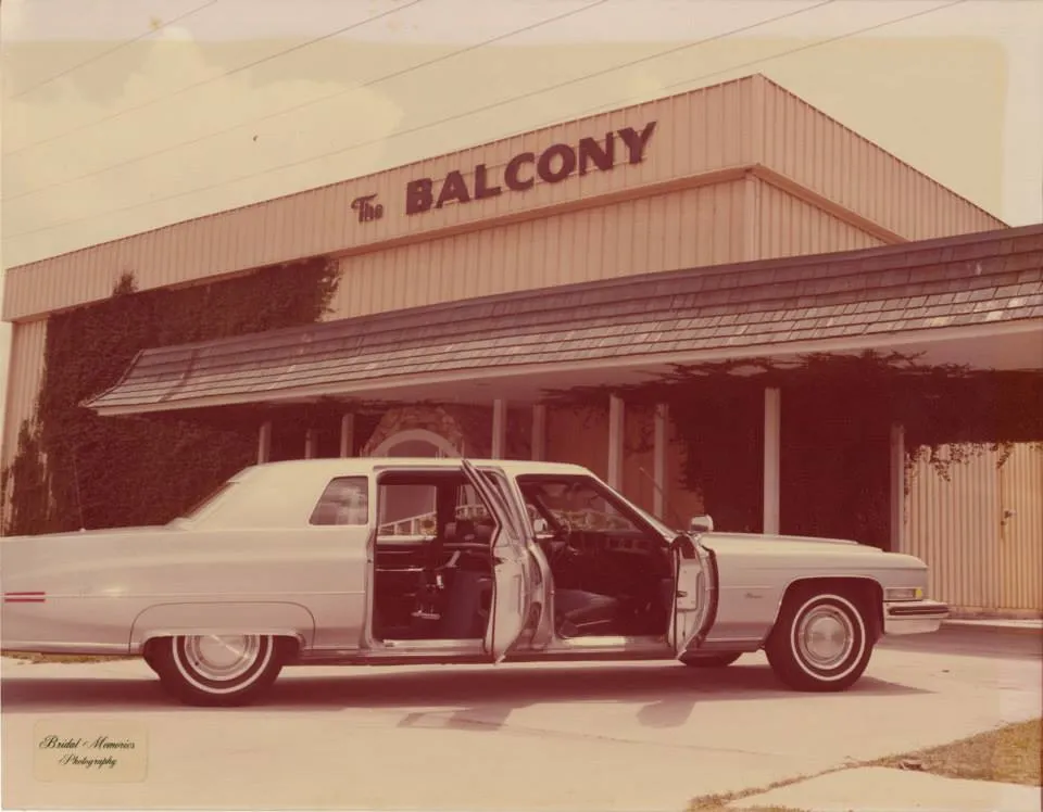 Vintage white car with open doors parked in front of a building with a sign reading The Balcony.