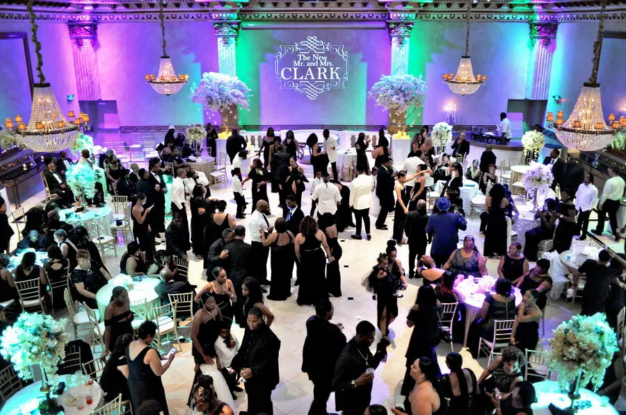 Elegant wedding reception with guests mingling and dancing under chandeliers and a wall projection reading 'The New Mr. and Mrs. Clark'.