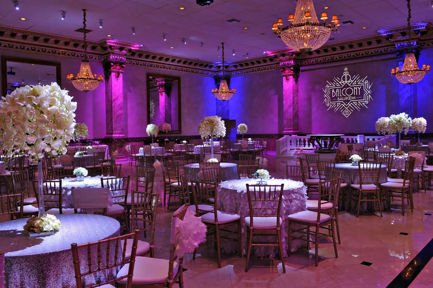 Elegant ballroom with round tables draped in silver and white linens, floral centerpieces, and ornate chandeliers under purple lighting.