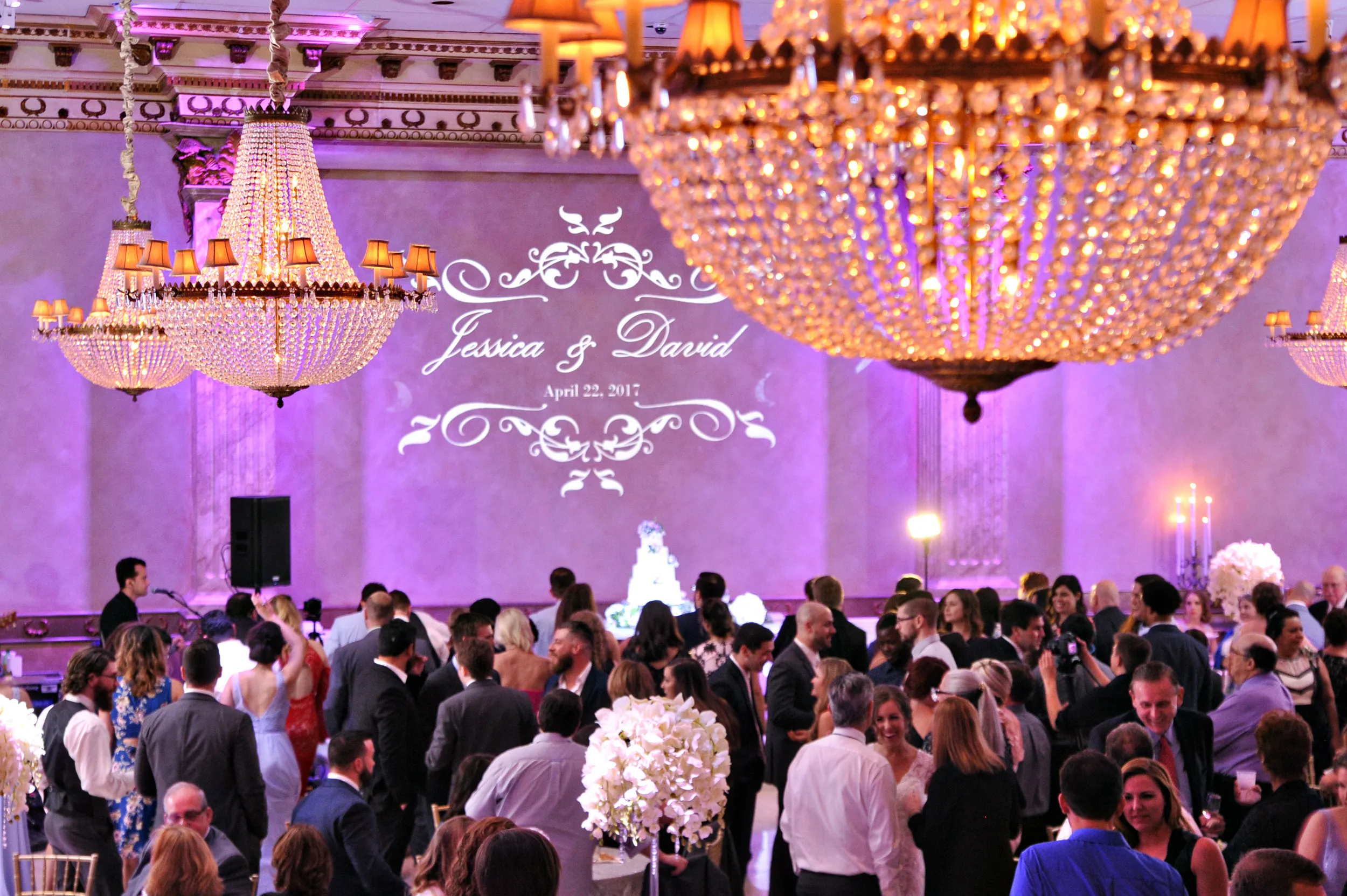 Wedding reception with guests mingling under large crystal chandeliers and a wall projection reading Jessica & David, April 22, 2017.