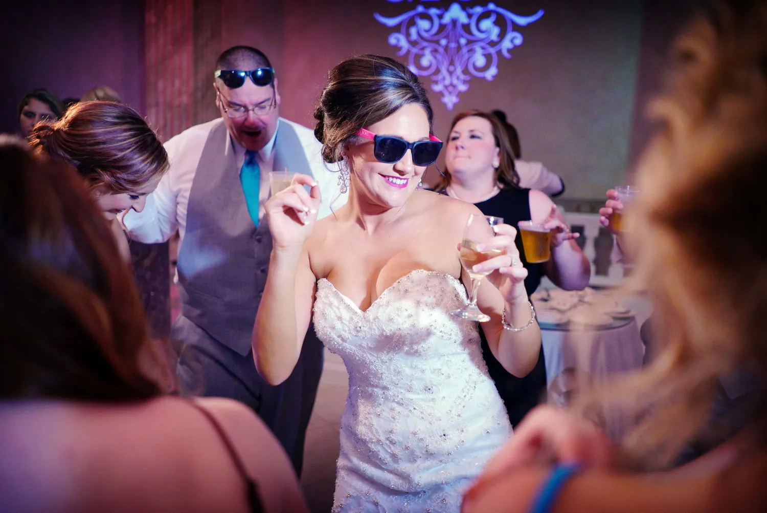 Bride wearing sunglasses dancing and holding a drink at a wedding reception with guests in the background.