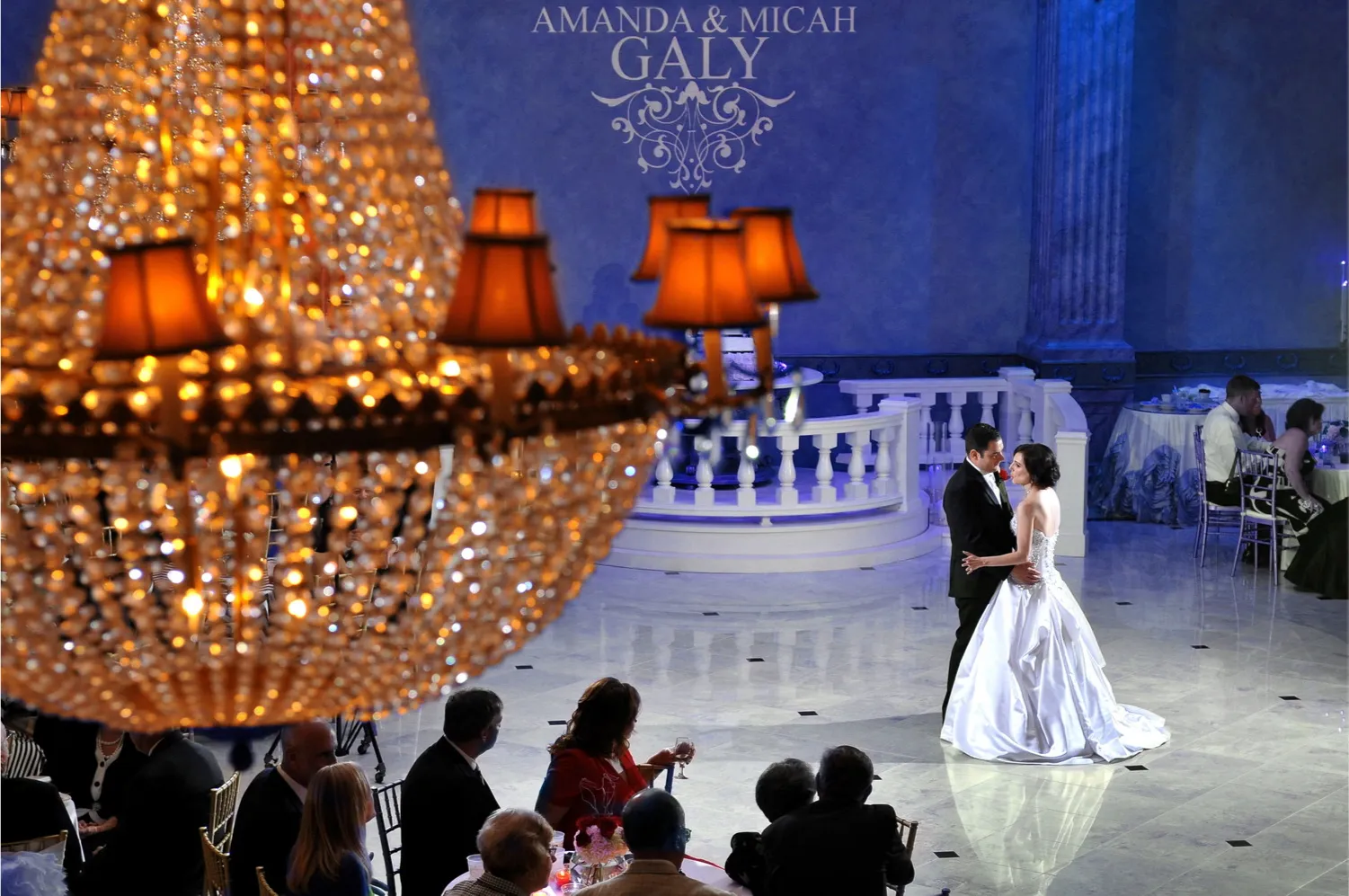 Bride and groom dancing on a marble floor in a grand hall with guests seated and a large chandelier in the foreground.