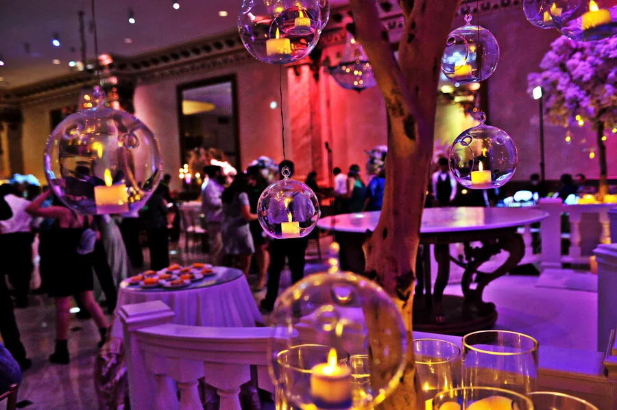 Indoor event with people socializing around tables and hanging glass globes with lit candles creating a warm, festive ambiance.