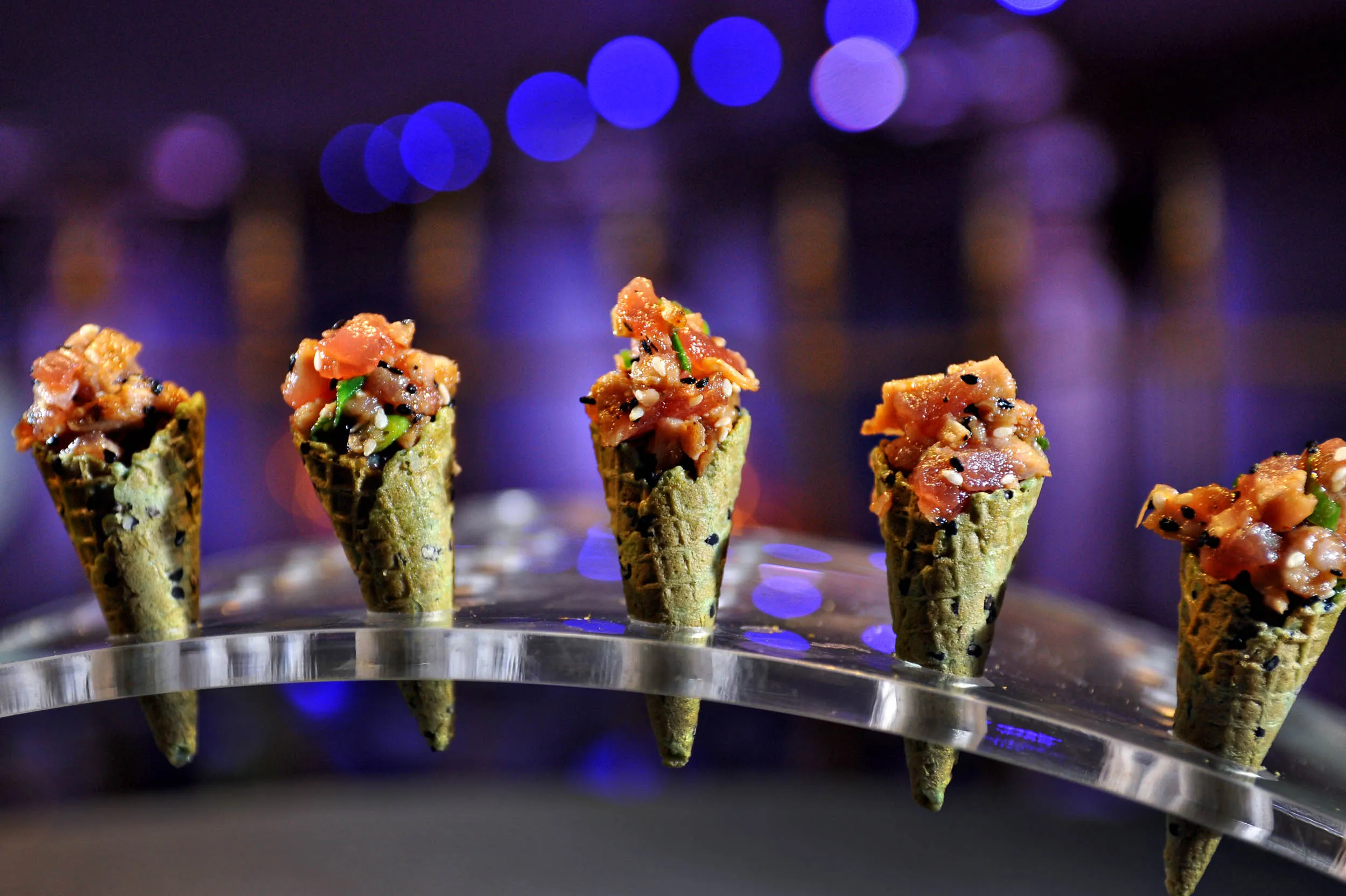 Five small green sesame waffle cones filled with diced raw fish topped with sesame seeds, arranged on a curved clear stand against a blurred blue-purple background.
