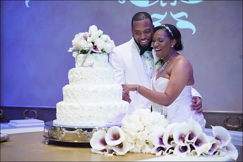 Bride and groom cutting a white wedding cake decorated with flowers, smiling together at their wedding reception.