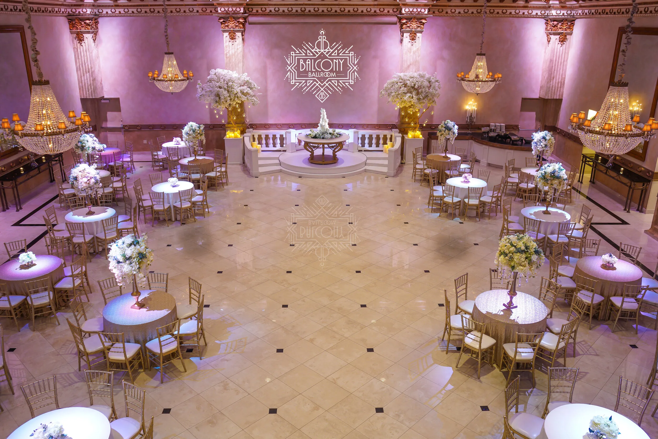 Elegant ballroom with round tables covered in beige tablecloths, floral centerpieces, gold chairs, crystal chandeliers, and a central stage with floral decor.