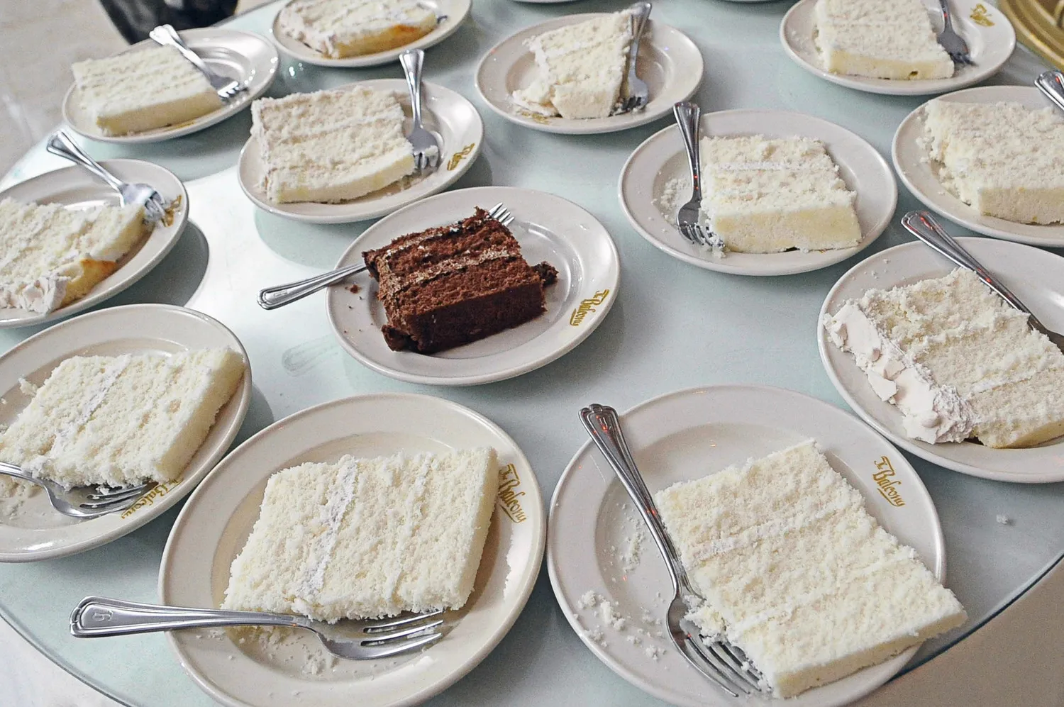 Multiple plates each with a slice of white cake with frosting and one plate with a slice of chocolate cake on a round glass table.