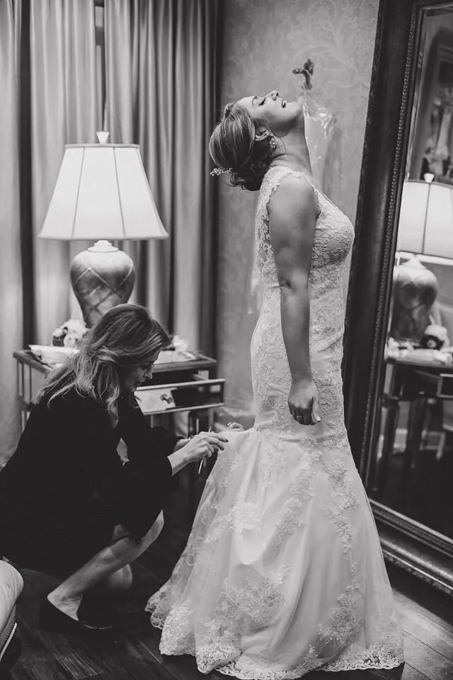 Black and white photo of a bride standing in a lace wedding dress while another woman adjusts the dress train.