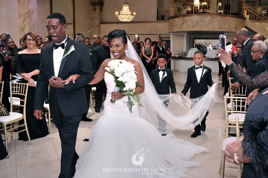 Bride in white wedding gown holding bouquet and smiling, escorted by man in black tuxedo, with young boys holding her veil and guests watching.