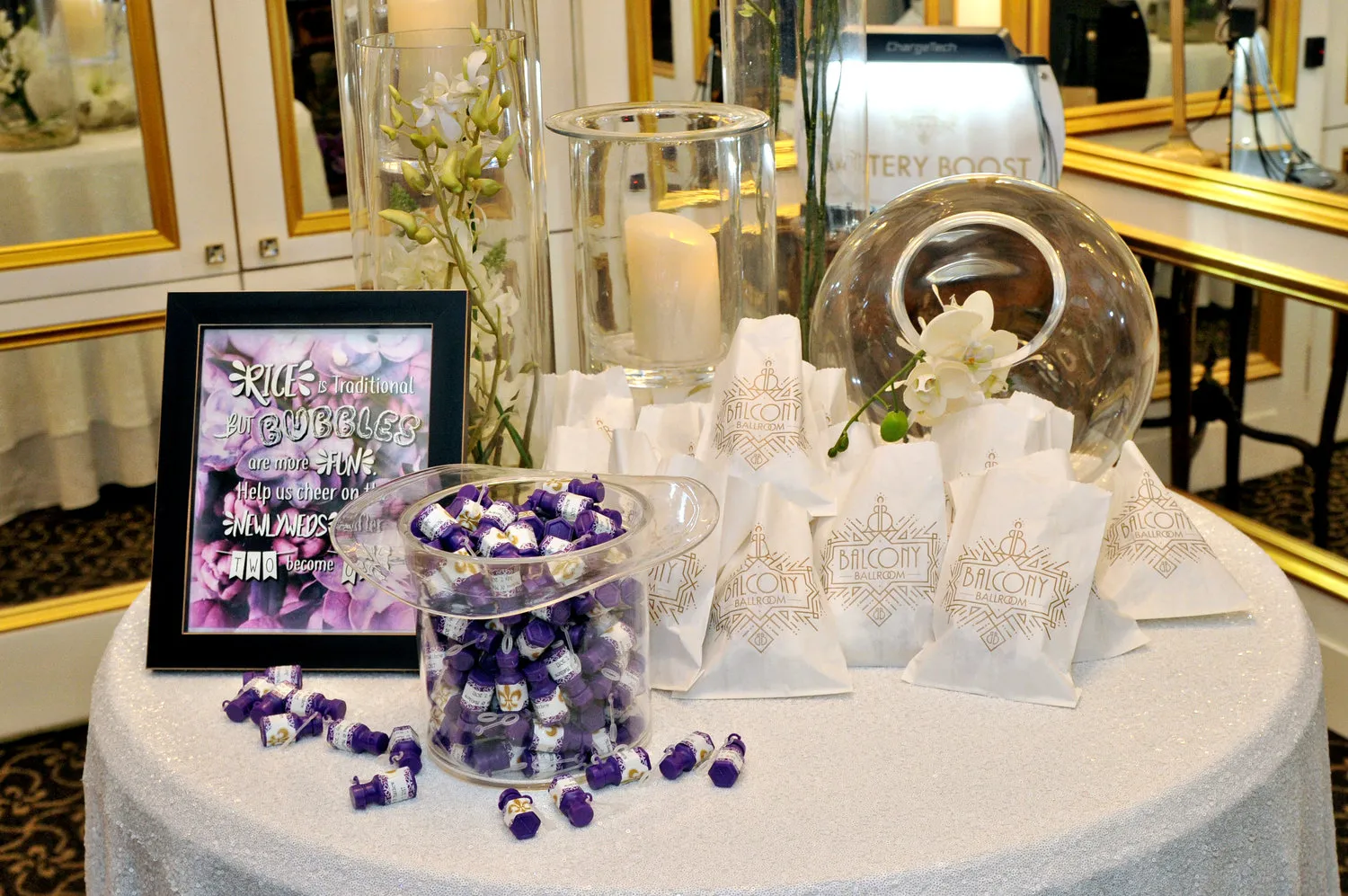 Wedding reception table with bags labeled 'Balcony Ballroom,' a glass bowl of purple bubbles, a candle in a glass holder, white flowers, and a framed sign encouraging guests to use bubbles instead of rice.