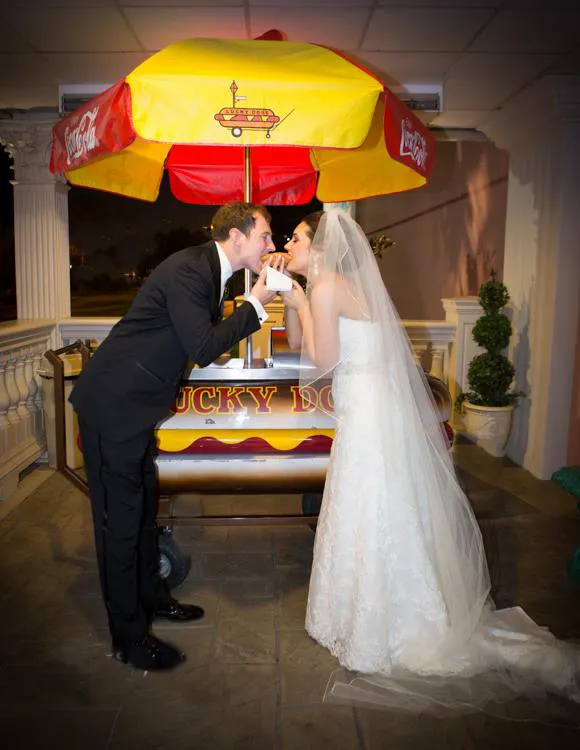 Bride and groom playfully feeding each other a hot dog under a yellow and red hot dog cart umbrella at night.