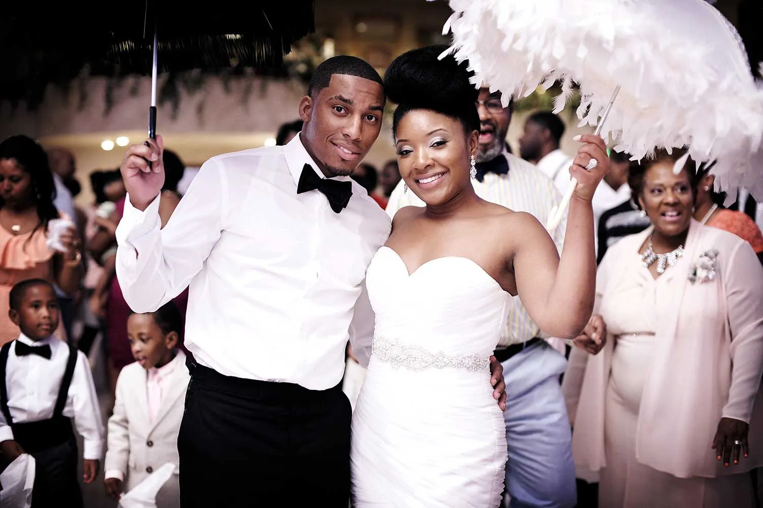 Smiling bride and groom dressed in wedding attire holding parasols, surrounded by guests at a celebration.