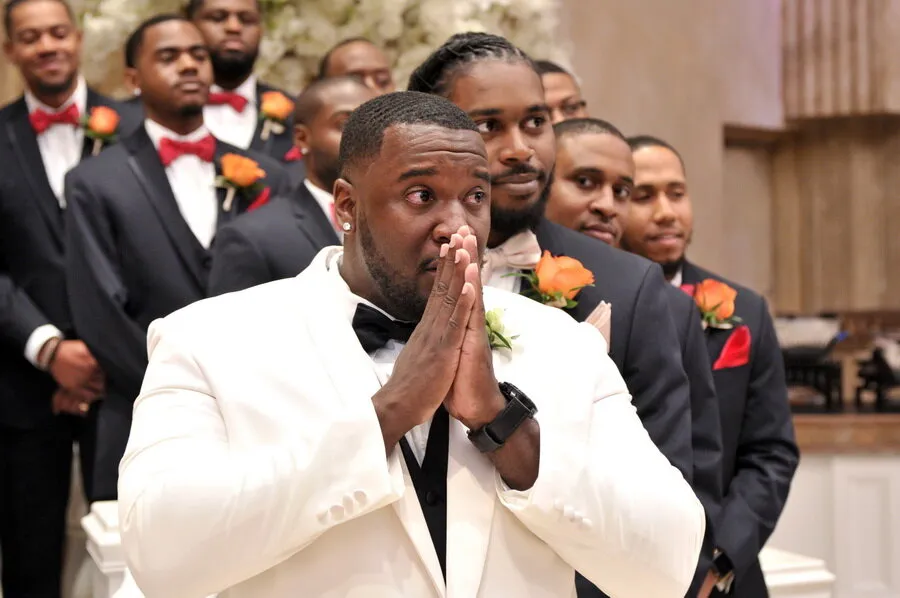 Groom in a white tuxedo looking emotional with hands clasped, standing in front of groomsmen in black suits with orange boutonnieres.
