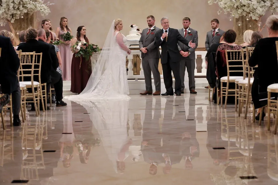 Bride and groom standing at the altar with an officiant speaking into a microphone, bridesmaids holding bouquets, and guests seated on gold chairs in a decorated indoor venue.