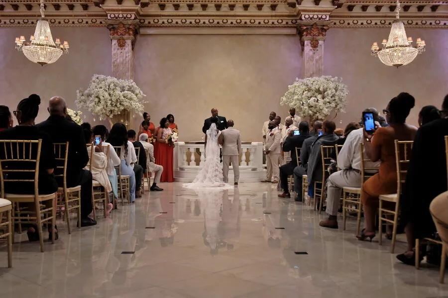 Bride and groom standing at an altar with an officiant during a wedding ceremony in an elegant hall with chandeliers and seated guests.