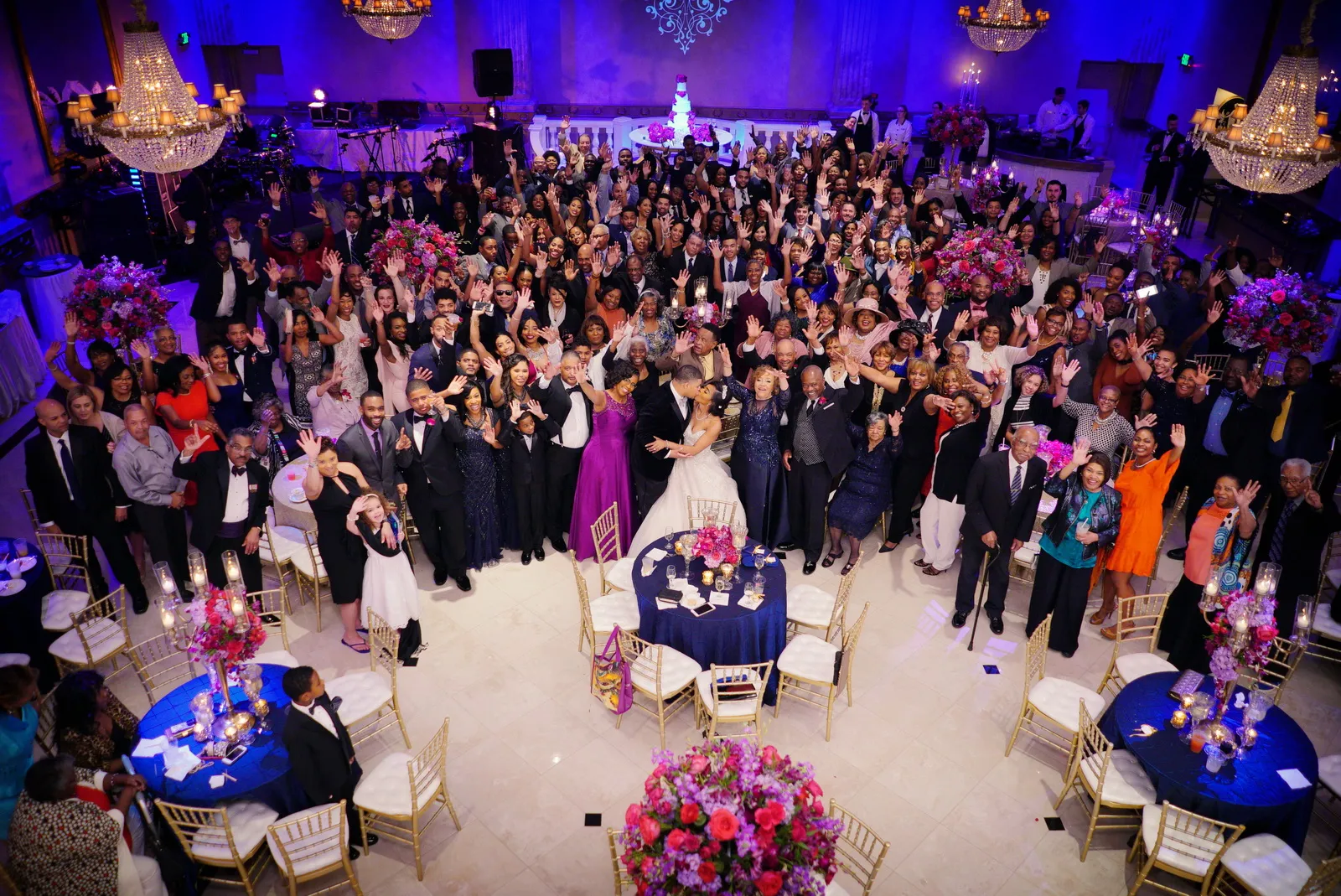 Large group of guests at a wedding reception, with bride and groom kissing in the center, many people smiling and waving.