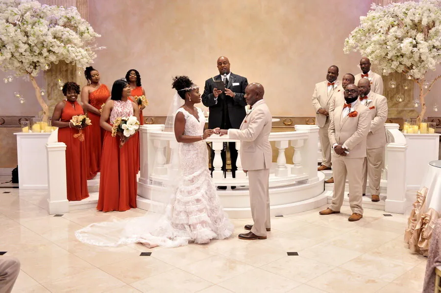 Bride and groom holding hands during wedding ceremony with officiant and wedding party dressed in red and beige.