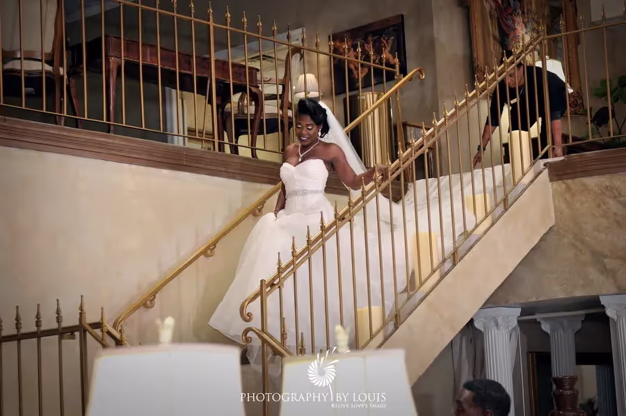 Bride in white dress with long train descending an indoor staircase with golden railings, smiling and holding the rail.