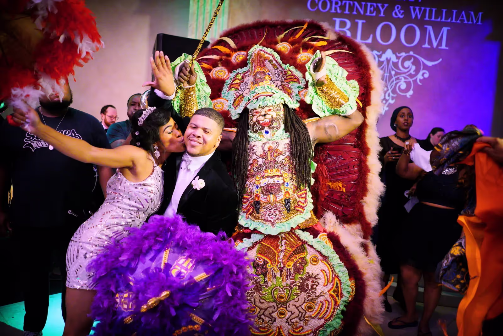 Woman in white dress kissing a smiling man in a tuxedo next to a person in a detailed, colorful Mardi Gras Indian costume with purple and red feathers at a lively celebration.