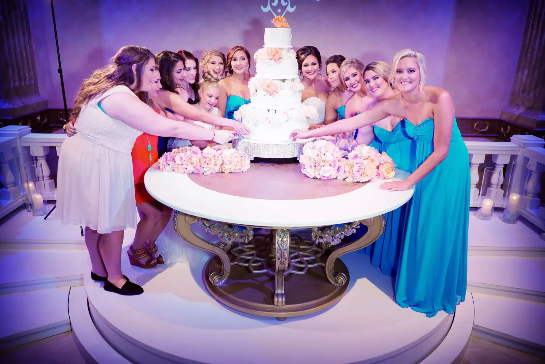 Bride and bridesmaids in blue dresses gathered around a multi-tiered white wedding cake on a decorated table with pink flower bouquets.