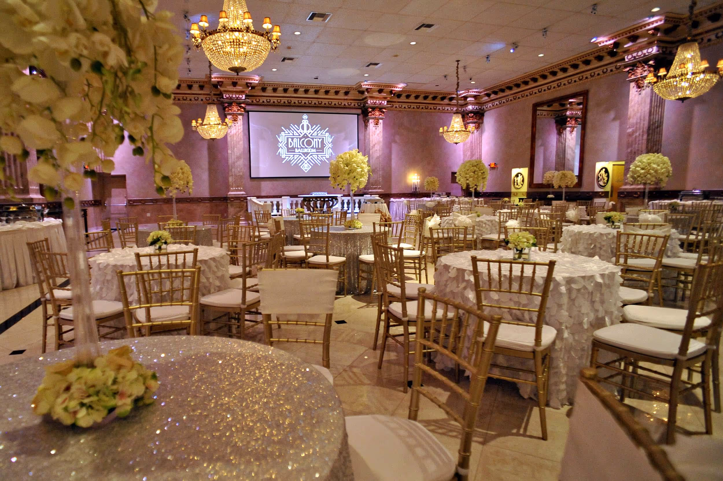 Elegant ballroom with round tables covered in white and silver tablecloths, gold chairs, floral centerpieces, and chandeliers overhead.