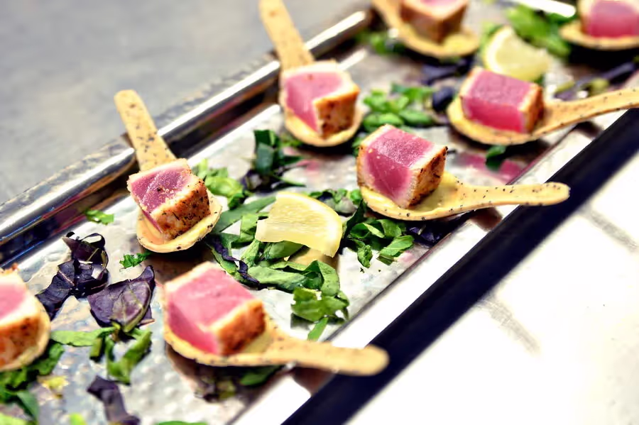 Seared tuna cubes served on wooden spoons over chopped greens and a lemon wedge on a metal tray.