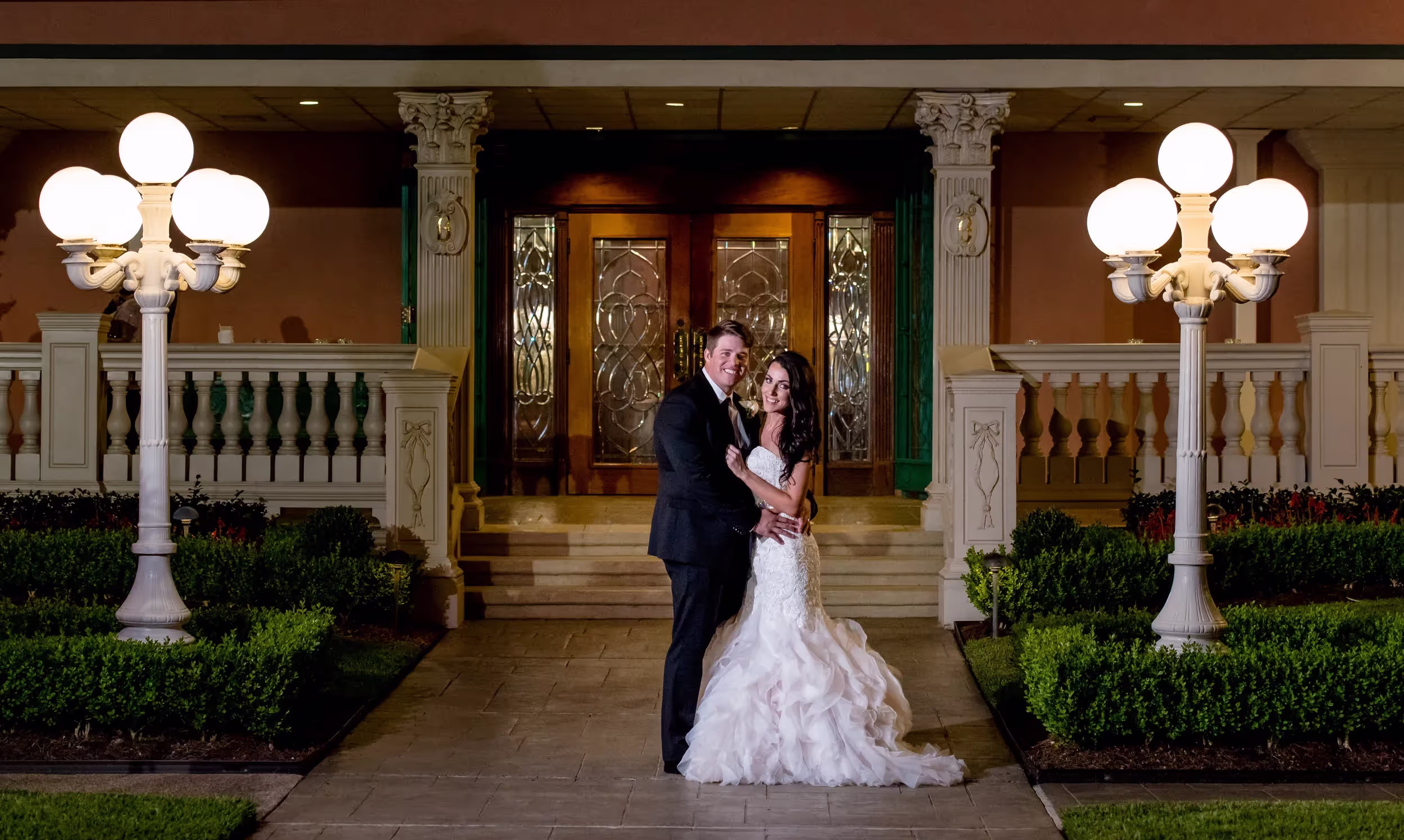 Bride in white wedding dress and groom in black suit standing and embracing in front of ornate double wooden doors at night.