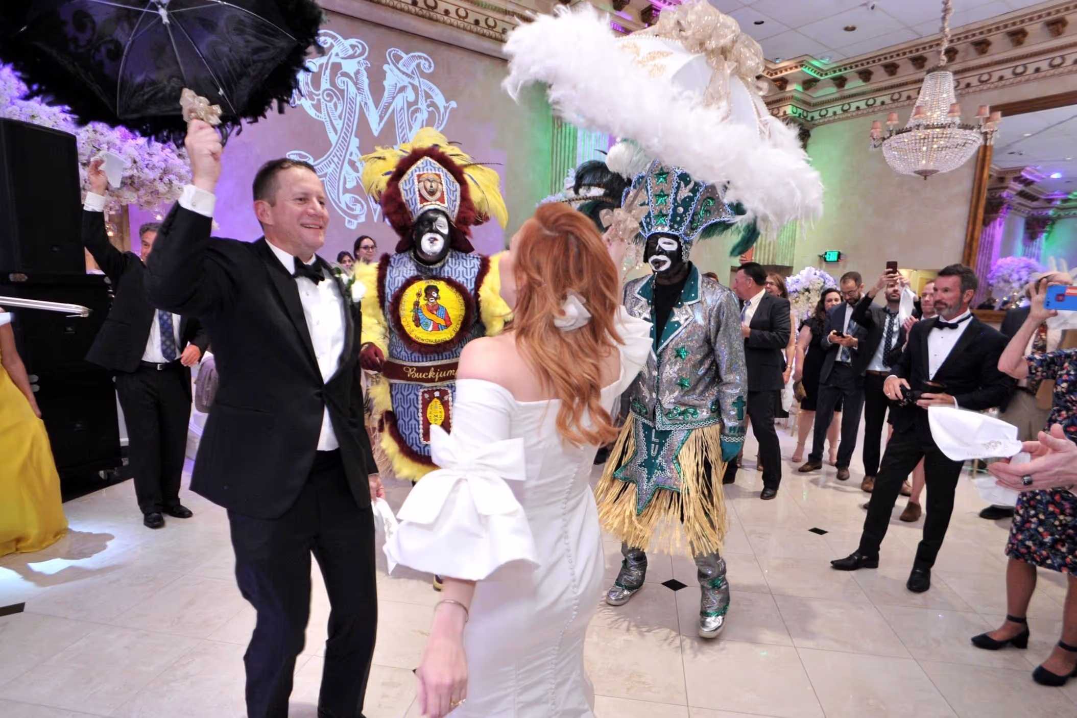 Bride and groom dancing surrounded by guests and two performers in colorful Mardi Gras Indian costumes at a wedding reception.