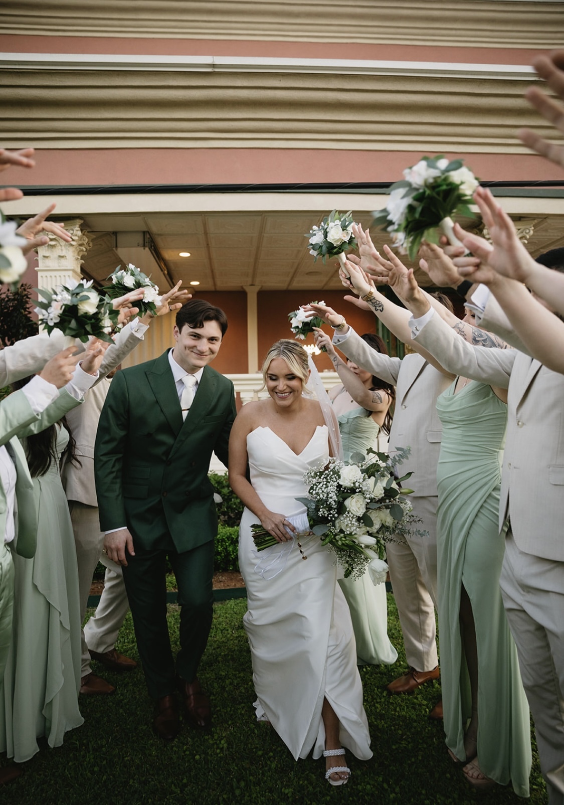 Bride and groom standing at the altar with an officiant speaking into a microphone, bridesmaids holding bouquets, and guests seated on gold chairs in a decorated indoor venue.
