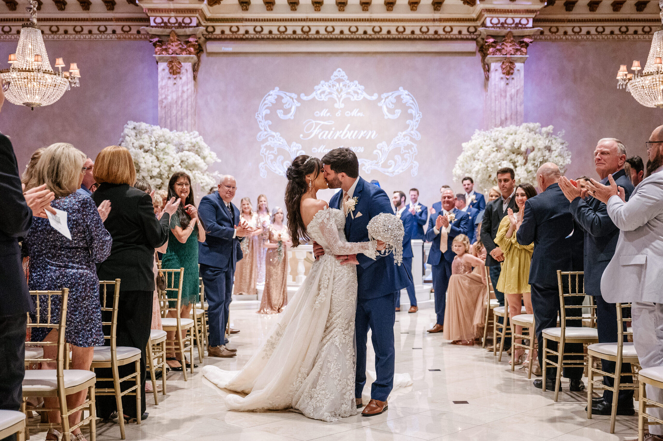 Bride and groom kissing indoors, each holding a feathered umbrella, with chandelier-lit background.