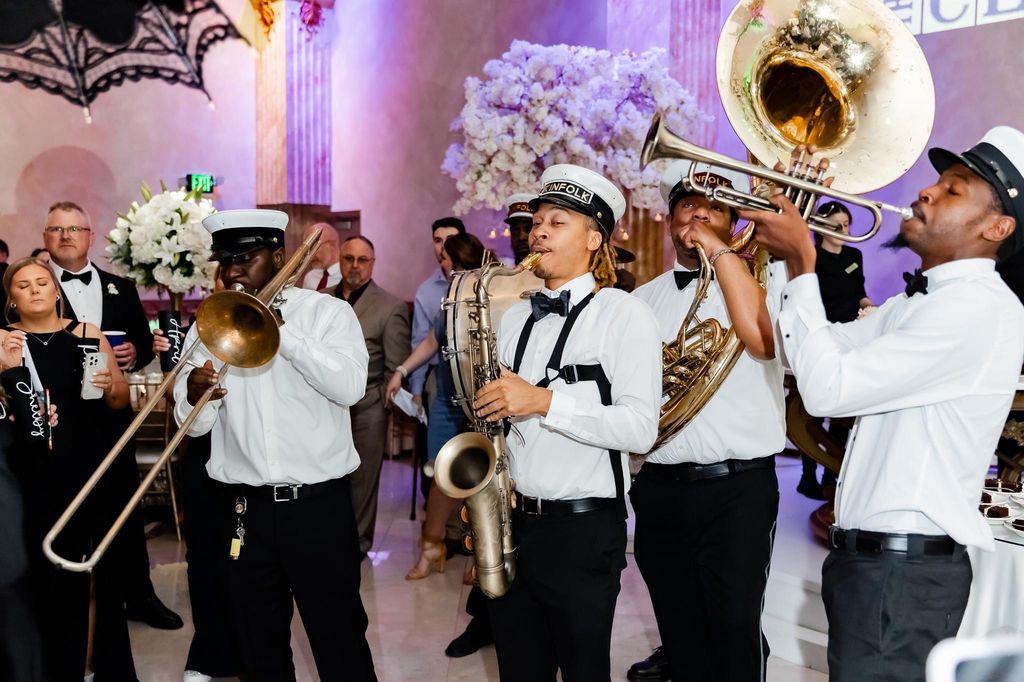 Large group of wedding guests dressed formally, gathered around a bride and groom kissing, all waving at the camera in a decorated reception hall.