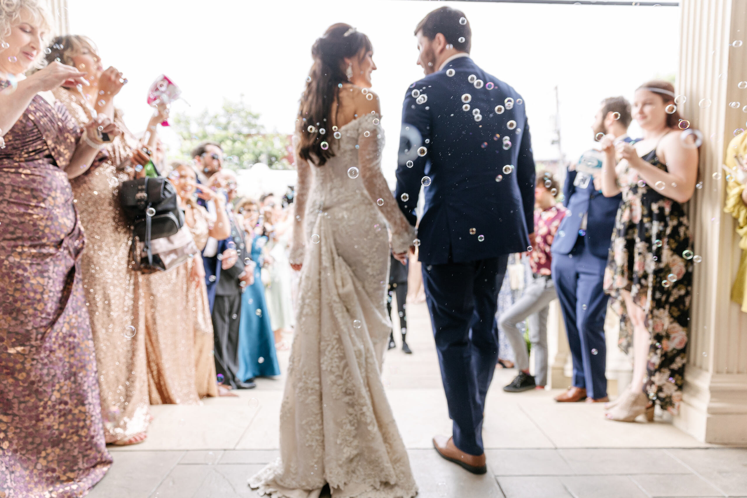 Bride in a white lace dress holding a cup and tipping a hat, surrounded by smiling wedding guests taking photos and watching.