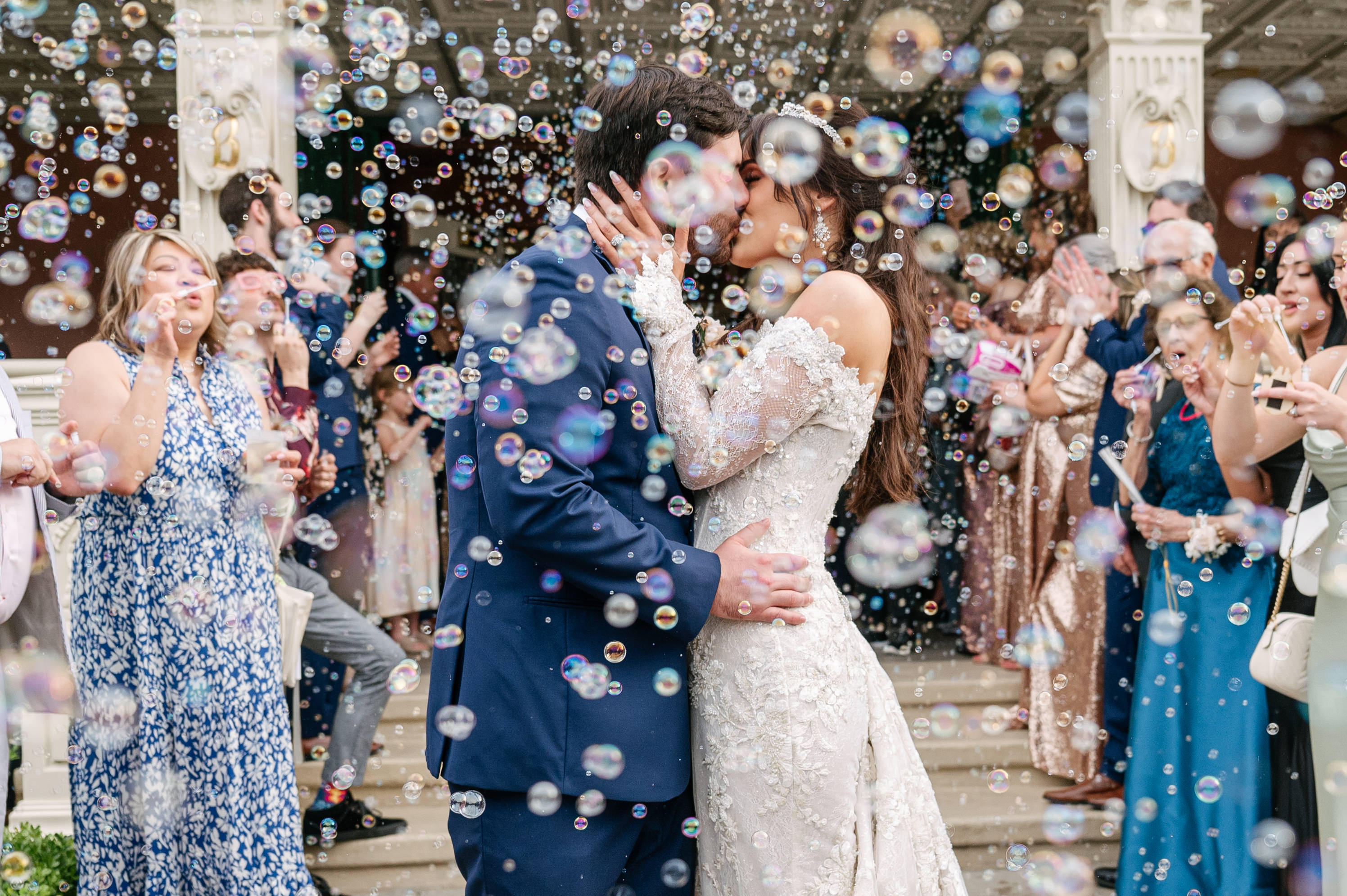 Large group of people at a wedding reception with the bride and groom kissing at the center, guests smiling and waving, surrounded by decorated tables and chandeliers.