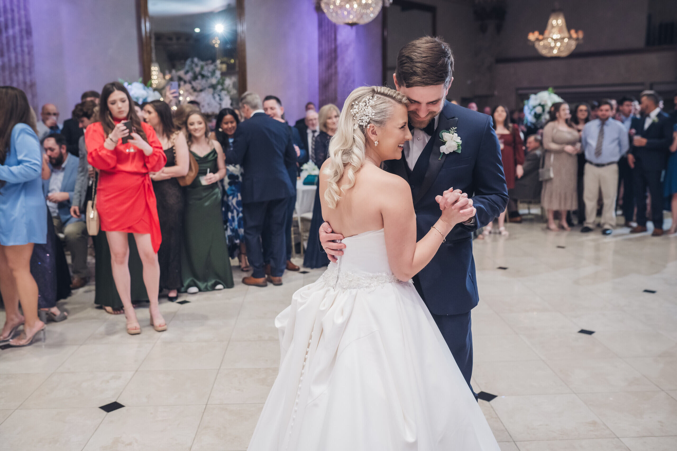 Bride wearing sunglasses dancing and holding a drink at a wedding reception with guests in the background.