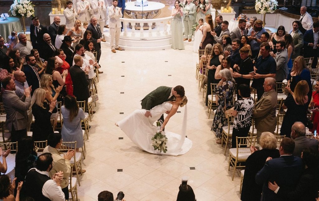Bride in white dress holding bouquet, smiling and walking arm-in-arm with a man in a black tuxedo, with two boys in tuxedos holding her veil behind them in a decorated indoor wedding venue.