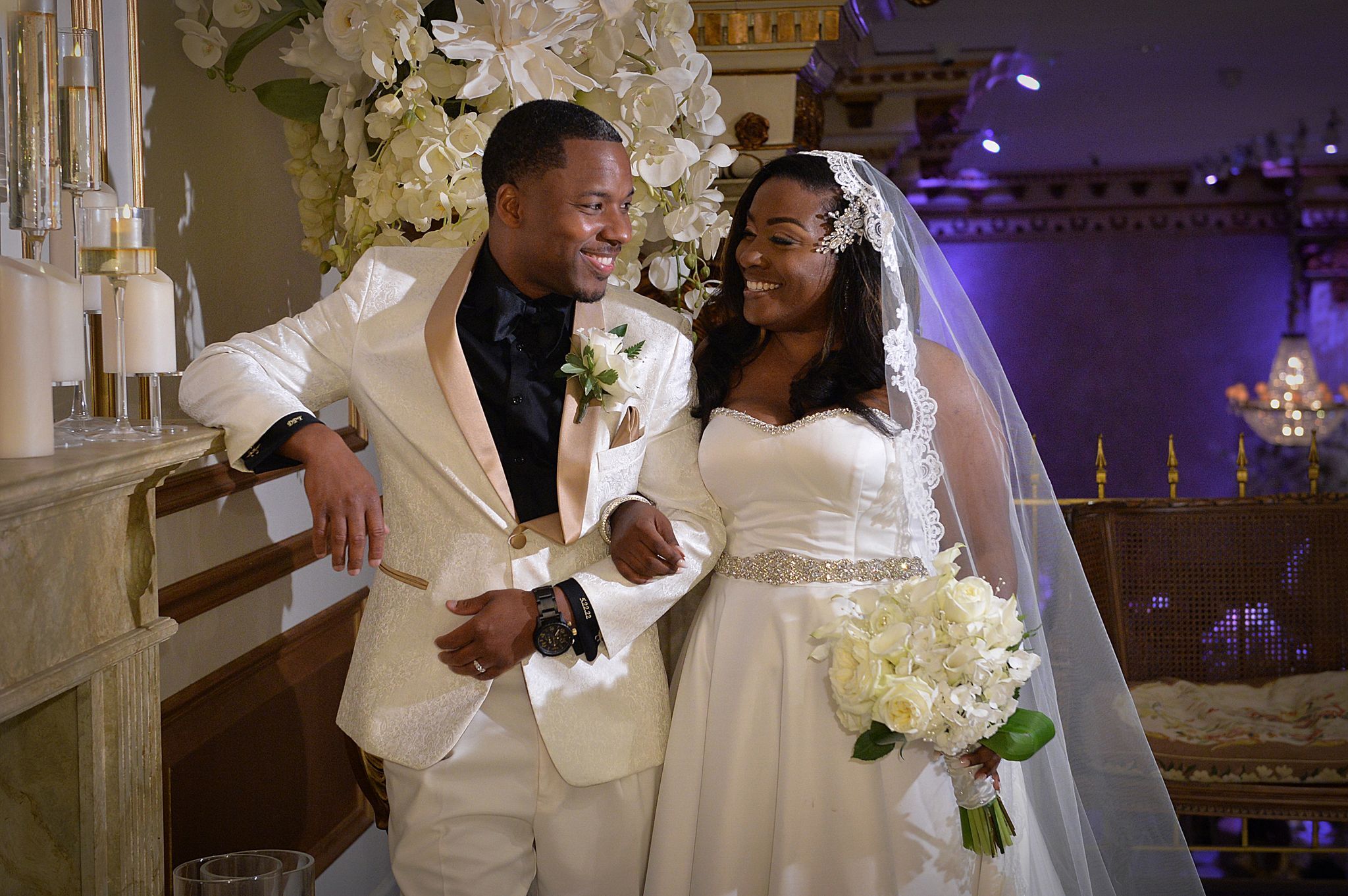 Bride and groom in wedding attire sitting at a round table, holding hands and kissing during reception.