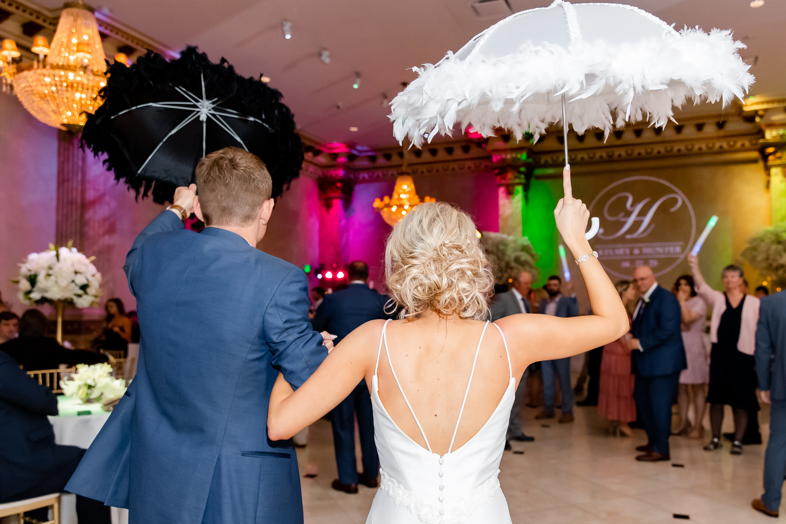 Large group of wedding guests dressed formally, gathered around a bride and groom kissing, all waving at the camera in a decorated reception hall.