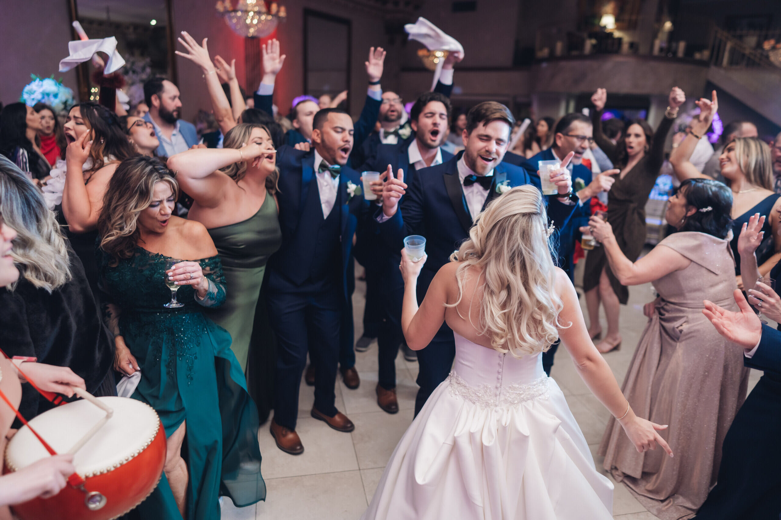 Bride in white wedding gown holding bouquet and smiling, escorted by man in black tuxedo, with young boys holding her veil and guests watching.