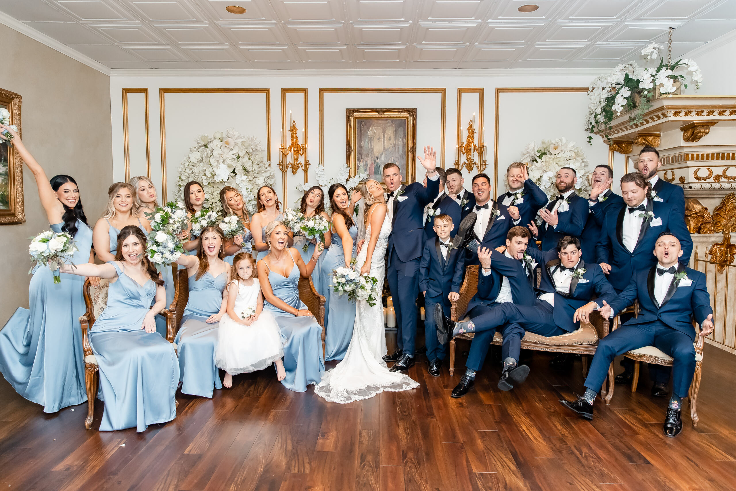 Large group of wedding guests gathered around the bride and groom who are kissing, all smiling and waving in a decorated reception hall.