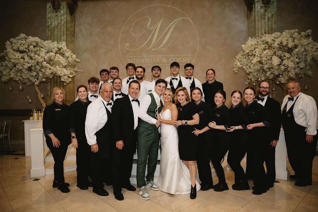 Bride in white wedding gown holding bouquet and smiling, escorted by man in black tuxedo, with young boys holding her veil and guests watching.