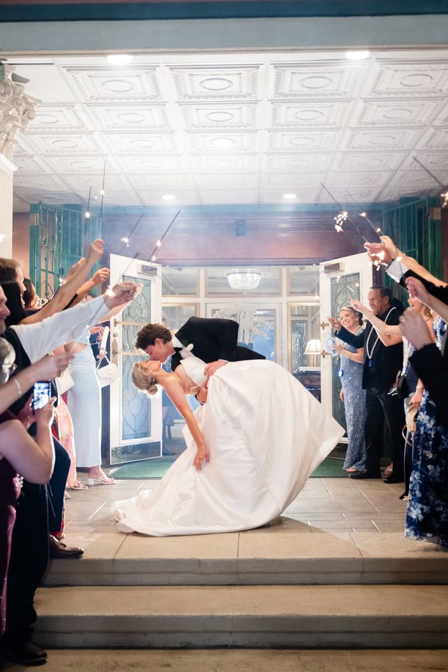 Bride and groom dancing on a marble floor in a grand hall with guests seated and a large chandelier in the foreground.