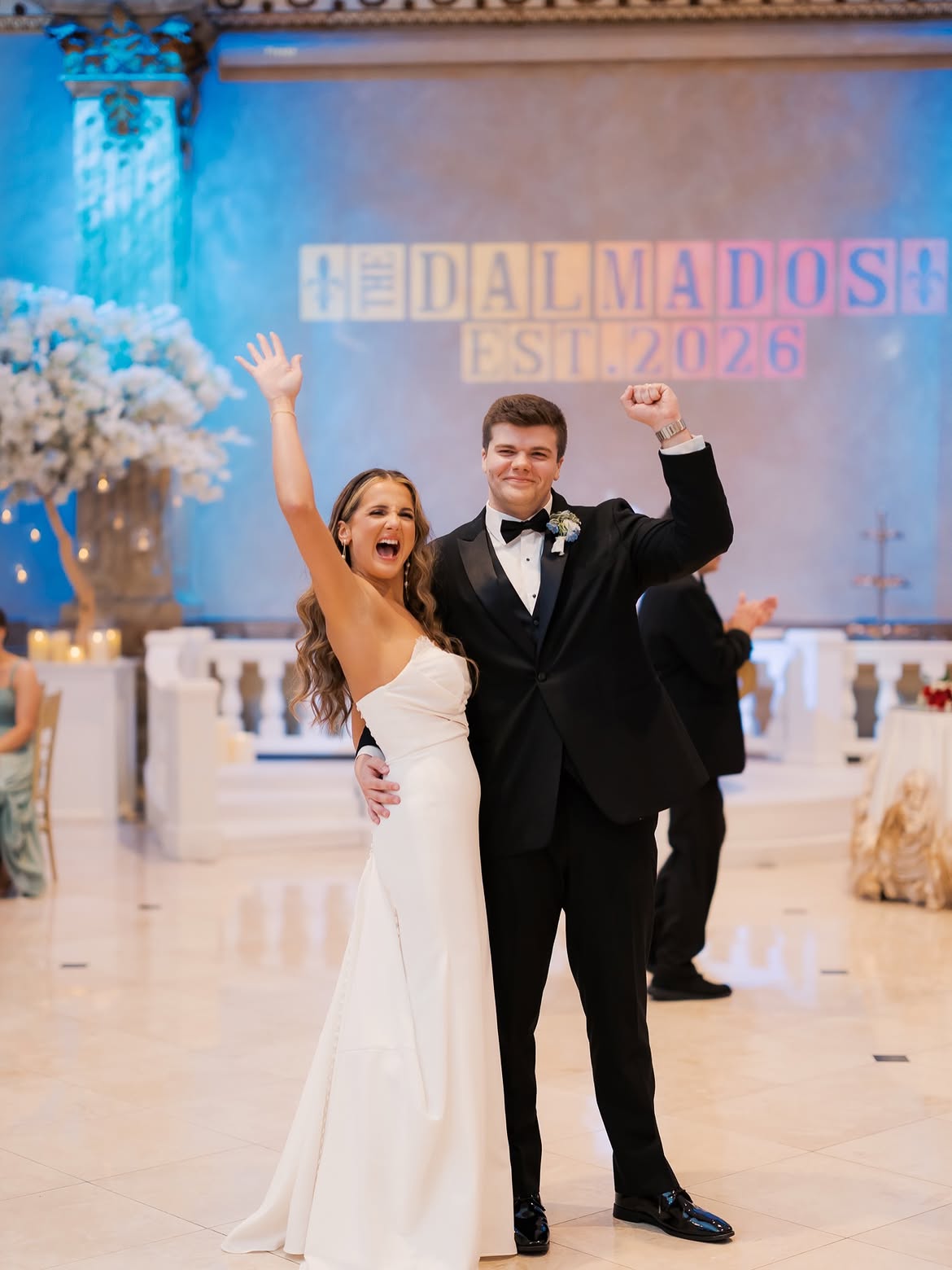 Bride in white wedding gown holding bouquet and smiling, escorted by man in black tuxedo, with young boys holding her veil and guests watching.