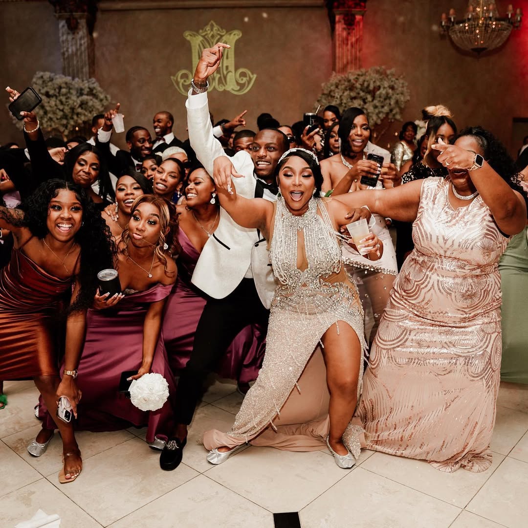 Bride and groom dancing on a marble floor in a grand hall with guests seated and a large chandelier in the foreground.