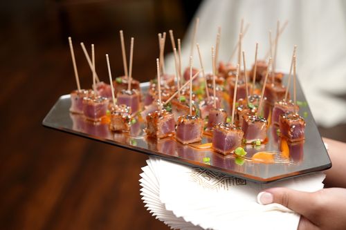 Five small green sesame waffle cones filled with diced raw fish topped with sesame seeds, arranged on a curved clear stand against a blurred blue-purple background.