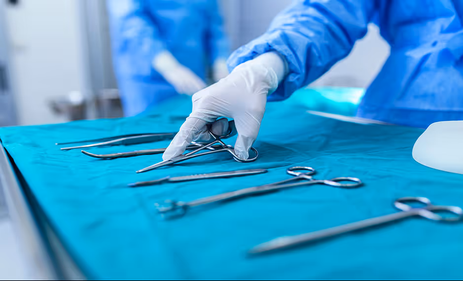 Gloved hand in surgical gown picking up a surgical clamp from a table with surgical tools.