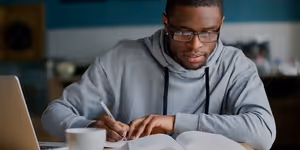 Man wearing glasses and gray hoodie writing notes from an open book at a table with a laptop.