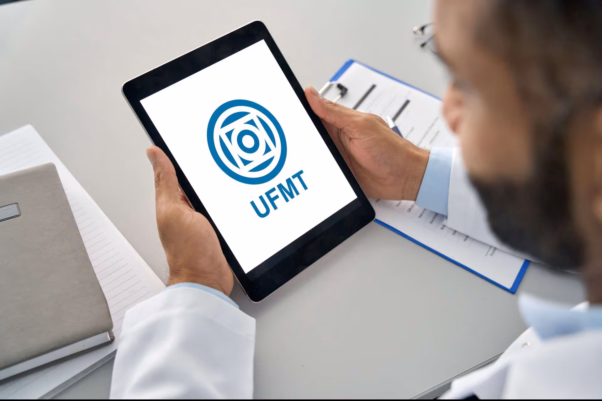 Person holding tablet displaying UFMT logo with documents and glasses on desk.
