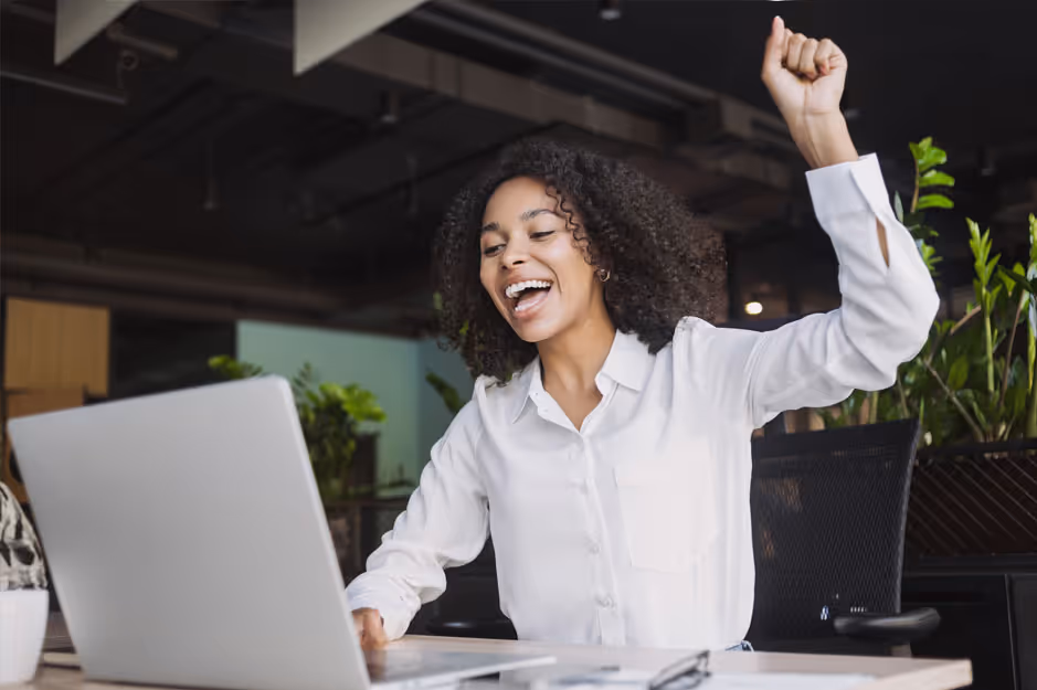 Woman in white shirt celebrating while using a laptop in a modern office.